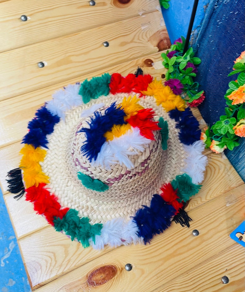 May include: A straw hat with a wide brim and colorful pom-poms around the edge. The pom-poms are in shades of red, yellow, blue, green, and white.