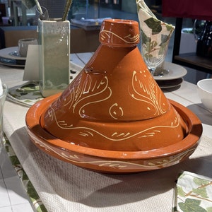 May include: A traditional Moroccan tagine with a brown clay pot and a cone-shaped lid. The lid and pot are decorated with a white floral design.