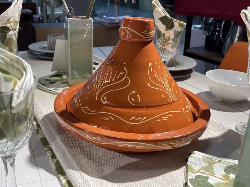 May include: A traditional Moroccan tagine with a brown clay pot and a cone-shaped lid. The lid and pot are decorated with a white floral design.