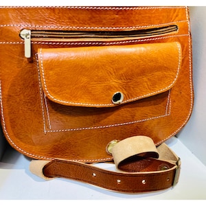 May include: A brown leather crossbody bag with a flap closure and a zippered pocket. The bag has a white stitching detail and a brown leather strap.