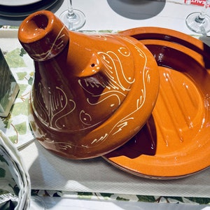 May include: A terracotta tagine with a lid featuring a white floral design. The tagine is a traditional Moroccan cooking pot.