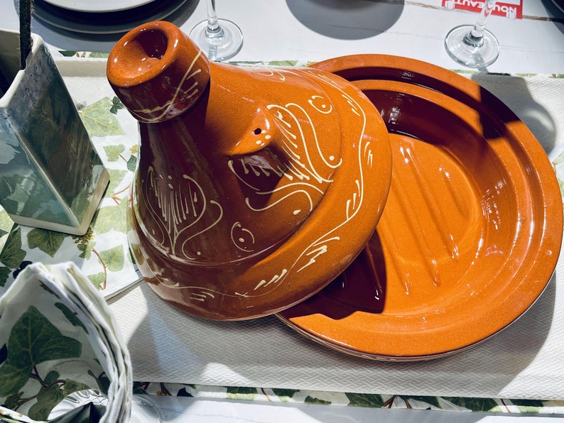 May include: A terracotta tagine with a lid featuring a white floral design. The tagine is a traditional Moroccan cooking pot.