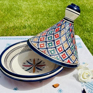 May include: A white ceramic tagine with a blue and white geometric pattern lid. The tagine is decorated with a colourful floral design.
