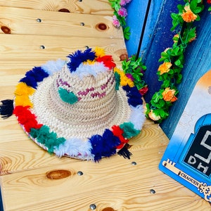May include: A straw hat with a wide brim and colorful pom-poms around the edge. The hat is made of woven straw and has a pink and white striped band.