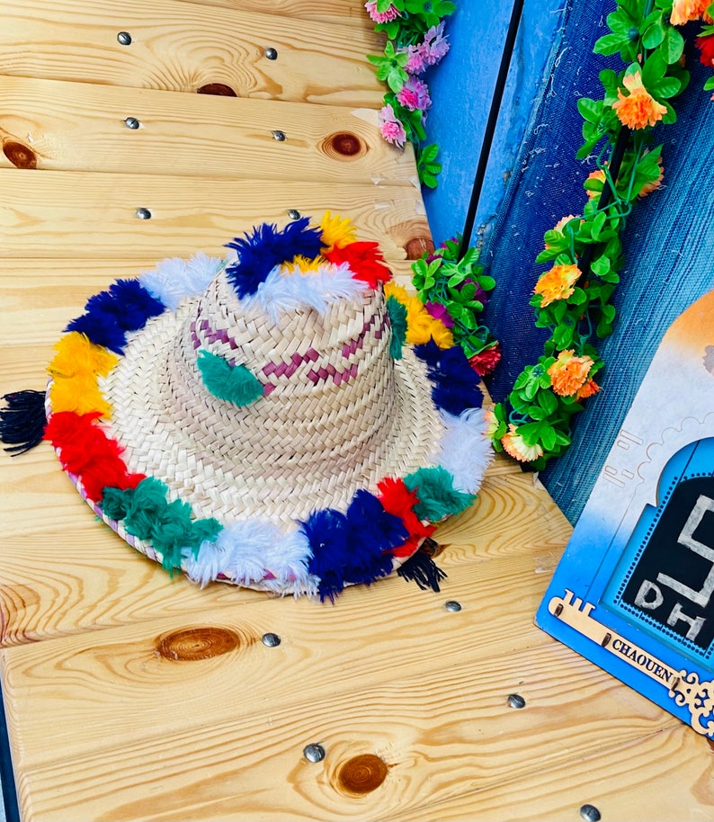 May include: A straw hat with a wide brim and colorful pom-poms around the edge. The hat is made of woven straw and has a pink and white striped band.