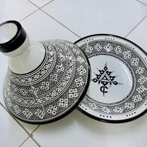 May include: A white ceramic tagine with a black and white geometric pattern. The lid has a black rim and the base has a black rim. The tagine is on a white tiled surface.