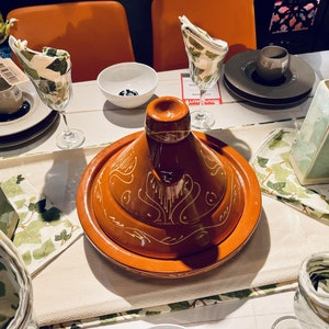 May include: A brown ceramic tagine with a white design, sitting on a white tablecloth with green and white napkins. The tagine is on a table with other dishes and glasses.