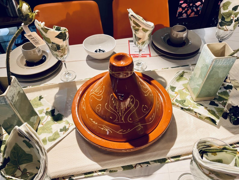 May include: A brown ceramic tagine with a white design, sitting on a white tablecloth with green and white napkins. The tagine is on a table with other dishes and glasses.