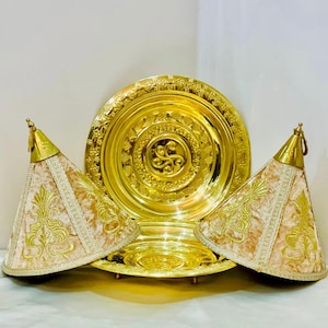 May include: A set of three gold-colored serving dishes with ornate designs. The two smaller dishes are covered with beige fabric with gold embroidery. The larger dish is round and has a raised center with a decorative pattern.