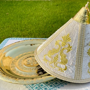 May include: A gold-colored serving tray with a matching lid. The lid is cream-colored with gold embroidery and trim. The tray and lid are on a white tablecloth with a teal and white pattern. The background is green grass.