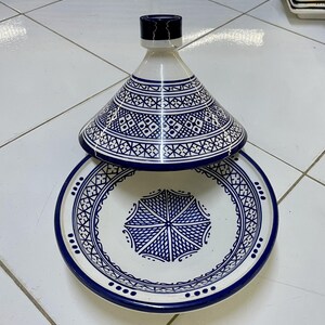 May include: A white ceramic tagine with a blue and white geometric pattern. The tagine has a round base and a conical lid.