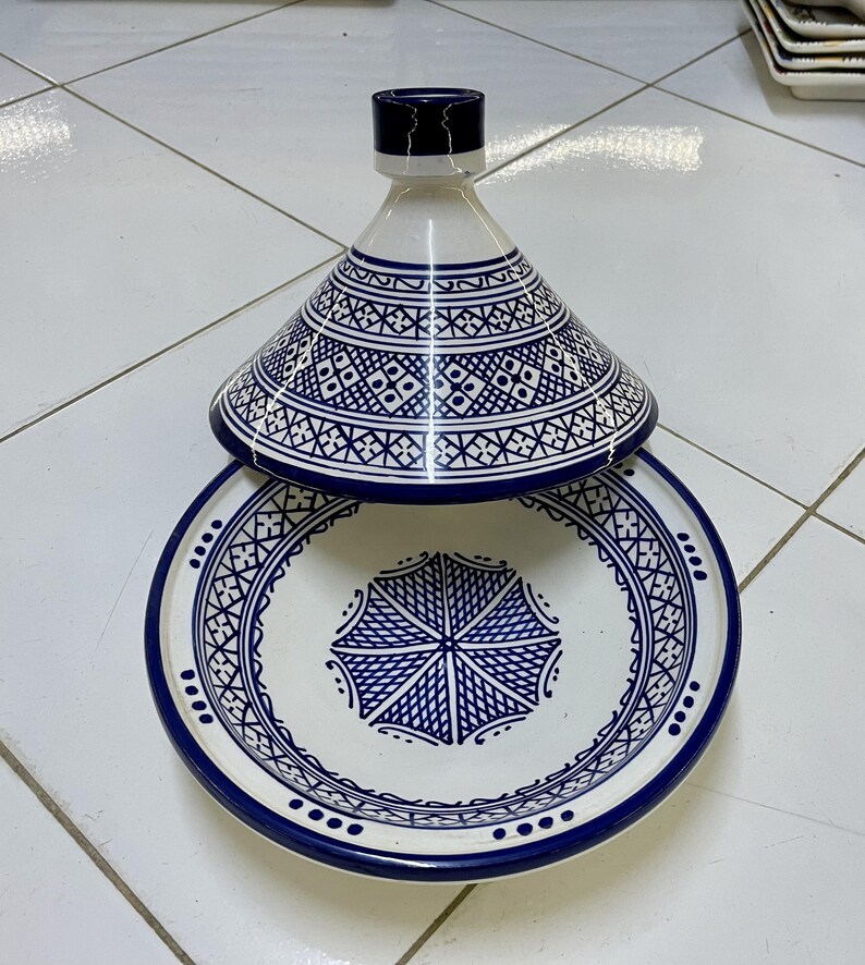 May include: A white ceramic tagine with a blue and white geometric pattern. The tagine has a round base and a conical lid.