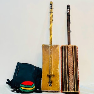 May include: Two traditional stringed musical instruments, both with wooden bodies and a single string. The instrument on the left has a rectangular body with a brown wooden surface and a yellow string. The instrument on the right has a cylindrical body with a brown wooden surface and a black string. Both instruments have a black carrying strap.