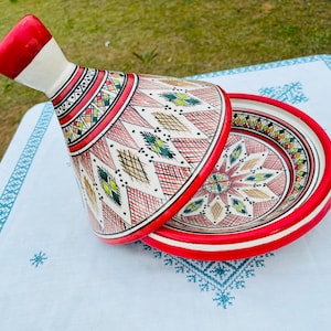 May include: A red and white ceramic tagine with a geometric pattern. The lid is open and the base is visible. The tagine is on a white tablecloth with a blue pattern.