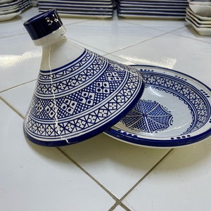 May include: A white ceramic tagine with a blue and white geometric pattern. The tagine is open and the lid is resting on the side of the dish.