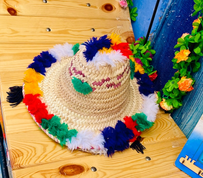 May include: A straw hat with a wide brim and colorful pom-poms around the edge. The hat is made of woven straw and has a pink and white striped band.