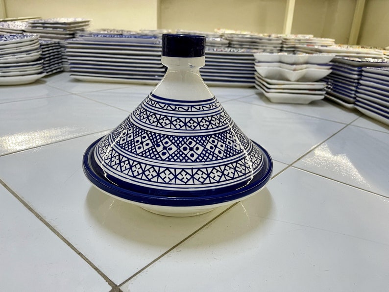 May include: A white ceramic tagine lid with a blue and white geometric pattern. The lid is round and has a pointed top. The lid is sitting on a white tiled surface.