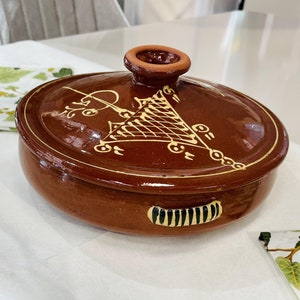 May include: A brown ceramic serving dish with a lid and a handle. The lid has a small knob on top and the dish is decorated with a yellow geometric design.