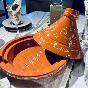 May include: A terracotta tagine with a white glaze and intricate designs. The lid is slightly ajar, revealing the interior of the pot. The tagine is on a white tablecloth with a green and white patterned napkin.