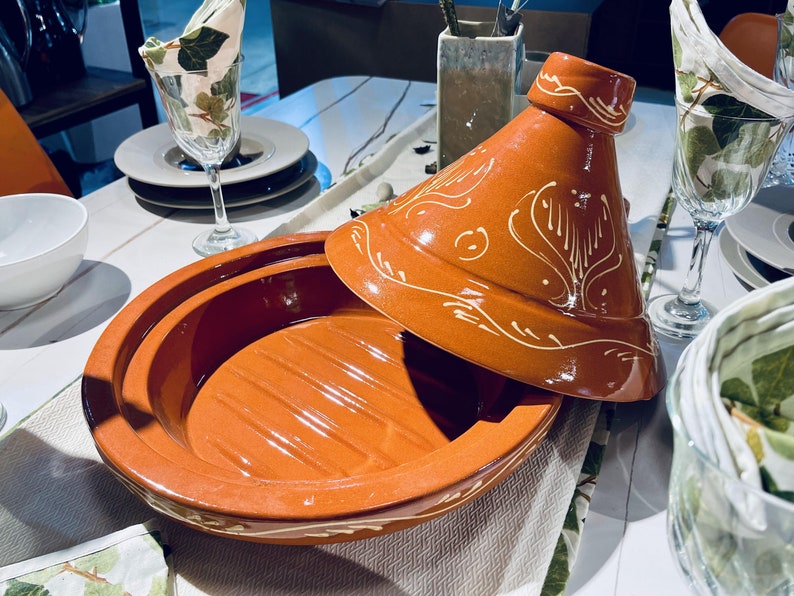 May include: A terracotta tagine with a white glaze and intricate designs. The lid is slightly ajar, revealing the interior of the pot. The tagine is on a white tablecloth with a green and white patterned napkin.