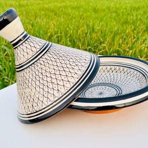 May include: A white ceramic tagine with a black and white geometric pattern. The tagine has a round base and a conical lid with a spout.