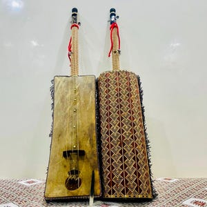 May include: Two traditional stringed musical instruments with wooden bodies and intricate designs. The instrument on the left has a rectangular body with a brown leather covering and a single string. The instrument on the right has a cylindrical body with a red and gold geometric pattern.