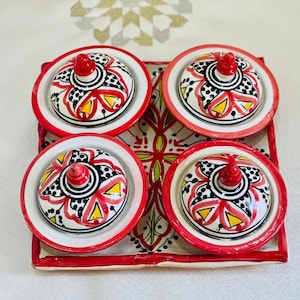 May include: A set of four small ceramic bowls with lids, each featuring a colorful floral design in red, black, white, and yellow. The bowls are arranged on a square tray with a red border and a white background.