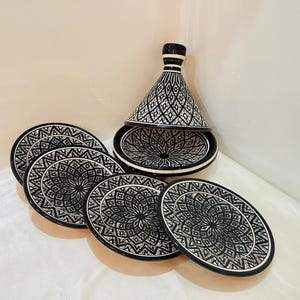 Pode incluir: Um conjunto de seis pratos de cerâmica preto e branco com intrincados desenhos geométricos. Os pratos estão dispostos em círculo em torno de uma tagine de cerâmica preto e branco com uma tampa cónica.