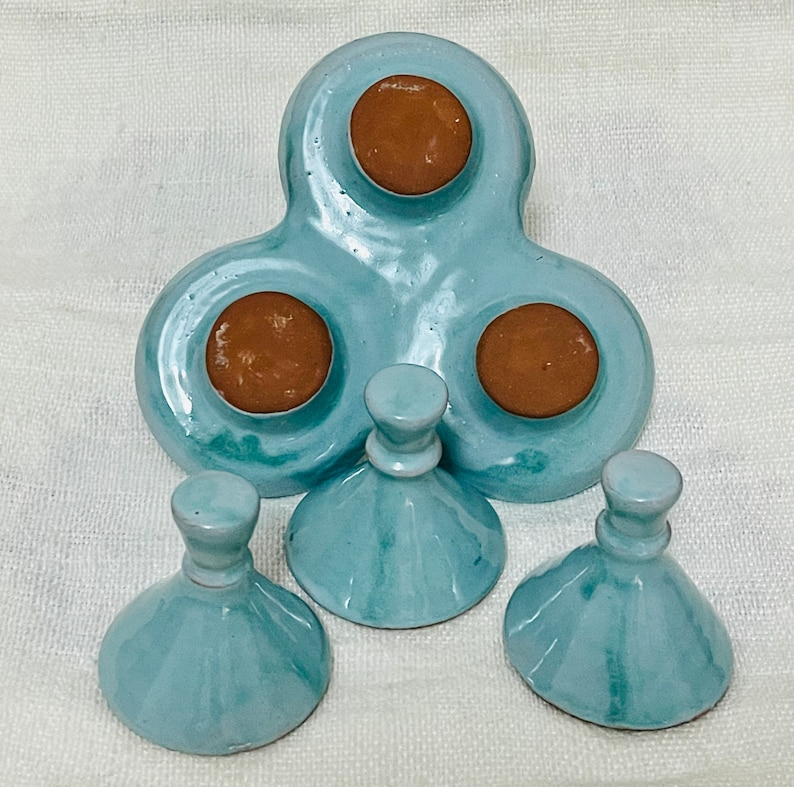 May include: A set of three teal blue ceramic condiment dishes with brown lids. The dishes are shaped like a clover leaf and have three individual compartments. The lids are round and have a smooth, glossy finish.