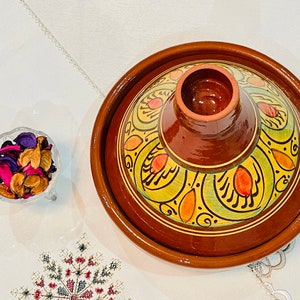 Puede incluir: Una tagine de cerámica marrón con un diseño floral amarillo, verde y naranja. La tagine tiene una tapa con una pequeña abertura en el centro.