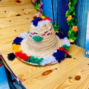 May include: A straw hat with a wide brim and colorful pom-poms around the edge. The hat is made of natural materials and has a woven texture.