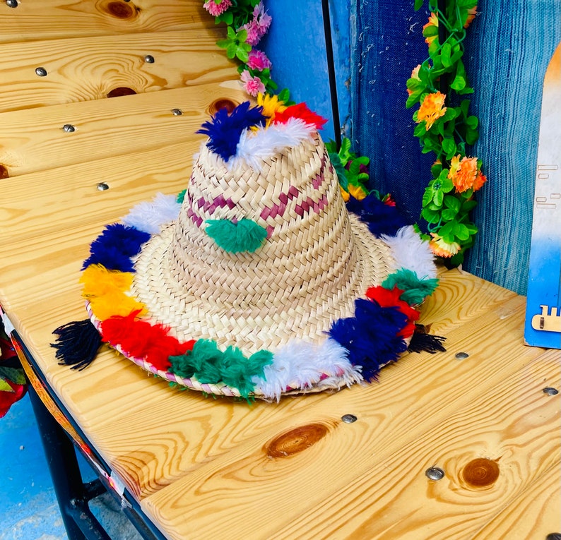 May include: A straw hat with a wide brim and colorful pom-poms around the edge. The hat is made of natural materials and has a woven texture.