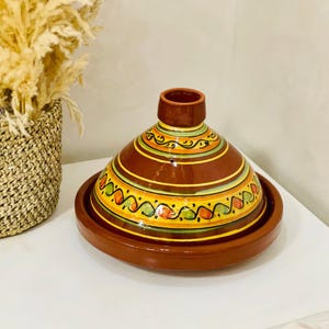 May include: A brown ceramic tagine pot with a conical lid and a circular base. The lid is decorated with colorful bands of yellow, green, and orange. The pot sits on a white surface.
