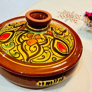 May include: A brown ceramic serving dish with a lid. The lid has a yellow, green, black, and red floral design. The dish is perfect for serving food.