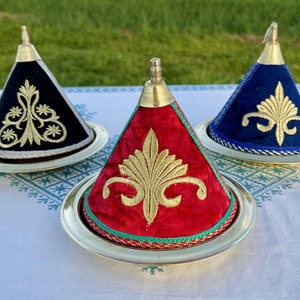 May include: Three decorative cone-shaped covers with gold embroidery on a white tablecloth. The covers are made of velvet fabric in black, blue, and red.