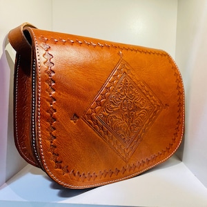May include: A brown leather satchel bag with a decorative embossed design. The bag has a flap closure and a shoulder strap.