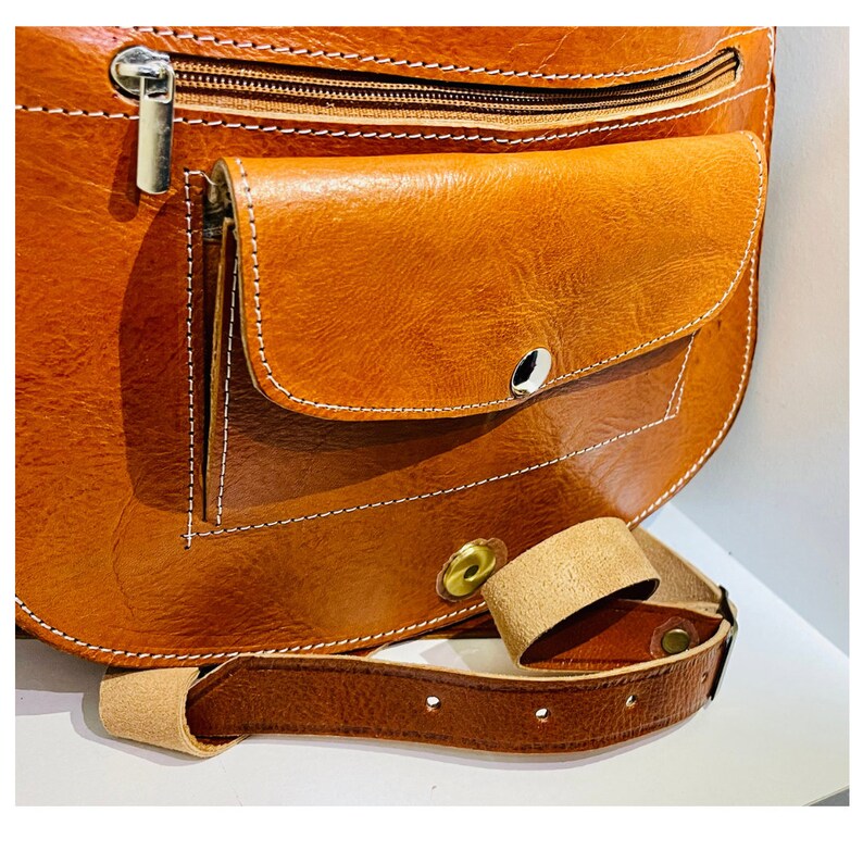 May include: A brown leather satchel bag with a flap closure and a zippered pocket. The bag has a brown leather strap with a metal buckle.