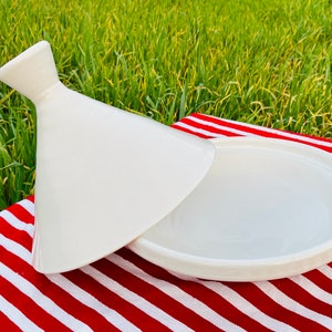 May include: White ceramic tagine with a conical lid. The lid is slightly ajar, revealing the round base of the tagine. The tagine is sitting on a red and white striped tablecloth.