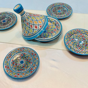 Pode incluir: Um conjunto de pratos de cerâmica pintados à mão e uma panela de tagine. Os pratos e a panela são decorados com intrincados padrões geométricos em tons de azul, vermelho, amarelo e verde. A panela de tagine tem uma tampa cónica.