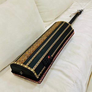 May include: A black stringed instrument with a long neck and a decorative body. The body features gold and brown geometric patterns and vertical stripes. The edges are trimmed with red, yellow, and black decorative trim. The instrument is resting on a white surface.