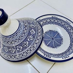 May include: A white ceramic tagine with a blue and white geometric pattern. The lid has a knob on top and the base has a wide, shallow bowl.
