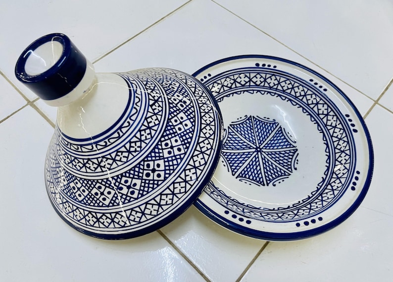 May include: A white ceramic tagine with a blue and white geometric pattern. The lid has a knob on top and the base has a wide, shallow bowl.