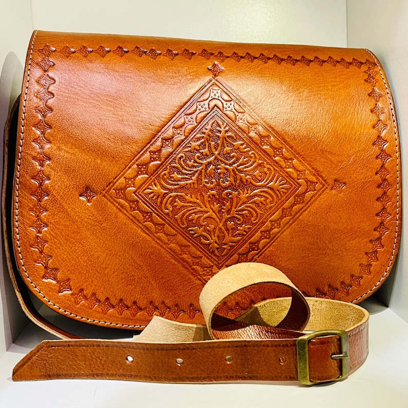 May include: A brown leather satchel bag with an intricate embossed design. The bag has a long adjustable strap and a flap closure.