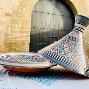 Puede incluir: Una tagine marroquí tradicional con una base blanca y patrones geométricos coloridos en rojo, verde, amarillo y azul. La tagine tiene una base redonda y una tapa cónica.