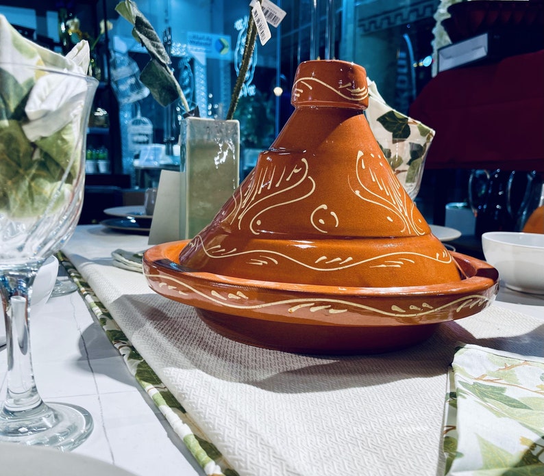 May include: A traditional Moroccan tagine cooking pot with a brown clay body and white decorative designs. The tagine is on a white tablecloth with a green and white patterned border.