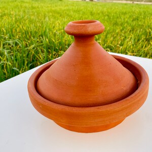 May include: A terracotta tagine cooking pot with a lid. The pot is round with a conical lid. The pot is sitting on a white surface.