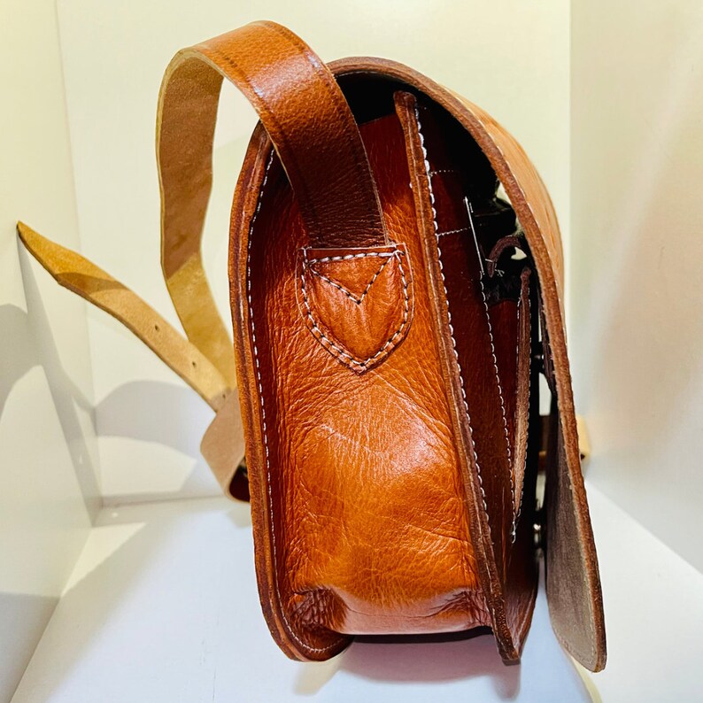 May include: A brown leather satchel bag with a long strap. The bag has a flap closure and a zippered pocket on the front.