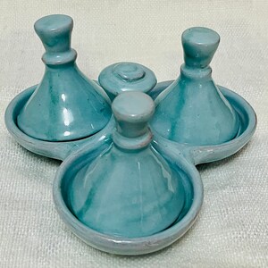 May include: A set of three blue ceramic condiment dishes with lids. The dishes are arranged in a circle on a white surface.