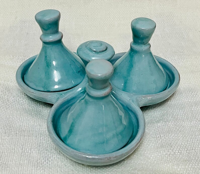 May include: A set of three blue ceramic condiment dishes with lids. The dishes are arranged in a circle on a white surface.