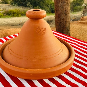 May include: A terracotta tagine lid with a round knob and a circular logo that reads "Terre Cuite" in a cursive font. The lid is sitting on a red and white striped tablecloth.